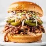 BBQ pulled pork sandwich with coleslaw and pickles on rustic wooden board.